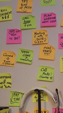 Vertical video. Woman placing colorful sticky notes with tasks and reminders on a wall while working late on her computer, selective focus. Time organization and planning