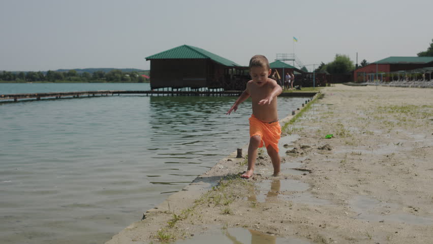 Young boy in bright orange swim shorts is playing along edge of lake. He runs excitedly on wet sandy shore, enjoying a fun day outdoors in the sun. In the background, the calm water and wooden piers.