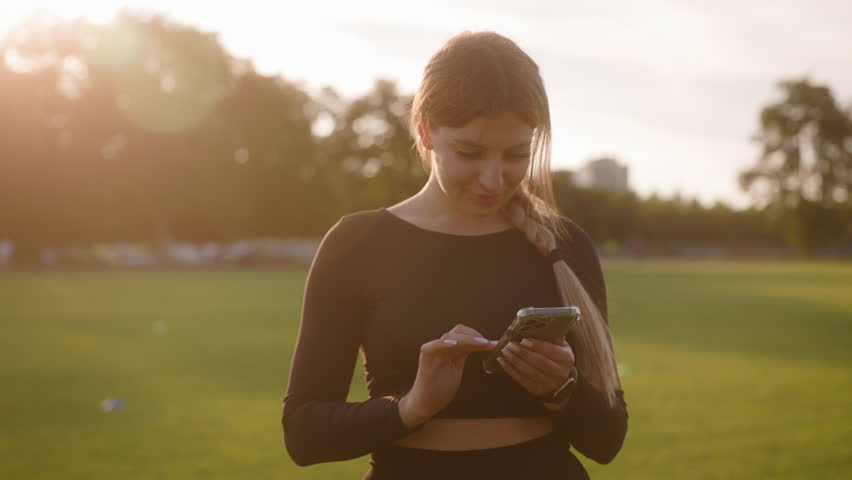 A young woman with a blonde braid stands outdoors on a green field, using her smartphone. She is wearing a black, long-sleeved sports top and is smiling as she interacts with her phone