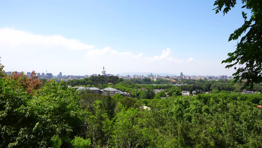Zoom in shot to Beihai White Tower seen from a lush green hilltop with Beijing skyline in the distance, China