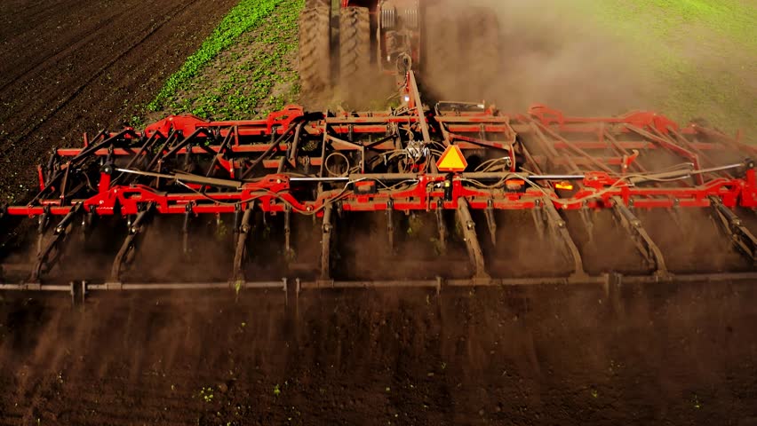 A large piece of agricultural machinery works diligently to till the soil on a farm during the spring season. Dust rises as the equipment readies the earth for the upcoming planting period.