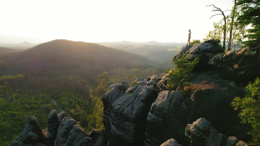 Hiker stands on top of rocky outcrop, overlooking a sunlit forest stretching into the horizon at dusk. Woman on a cliff edge, gazing into the vast valley below with a sense of wonder and discovery.