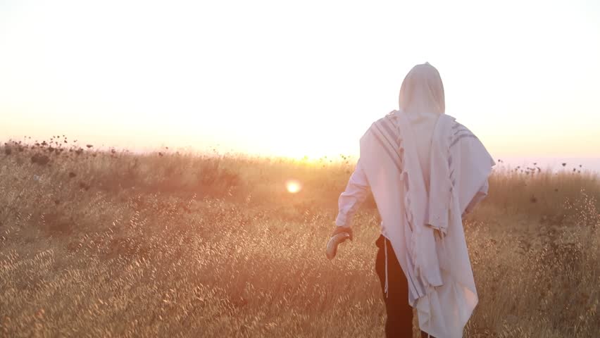 Jewish man in a traditional tallit prayer shawl blowing the ram's horn shofar, in the field against sunrise sky on Rosh HaShana and Yom Kippurim 