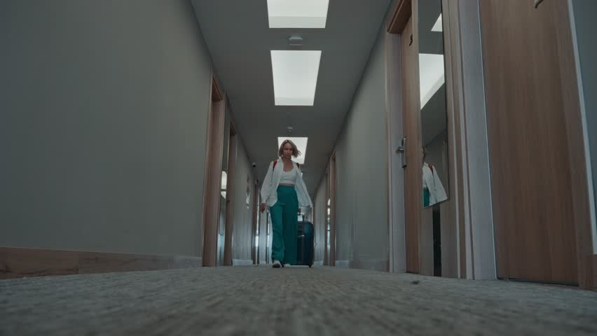 A slender woman walks down the hotel corridor with a suitcase. The woman is about to check into her room.