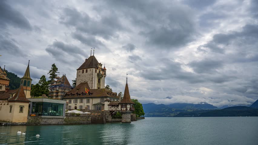 Timelapse of heavy clouds moving over Castle Oberhofen on the shore of Lake Thun in Switzerland, featuring medieval architecture with cloudy skies and mountain views in the background. 4K UHD video.