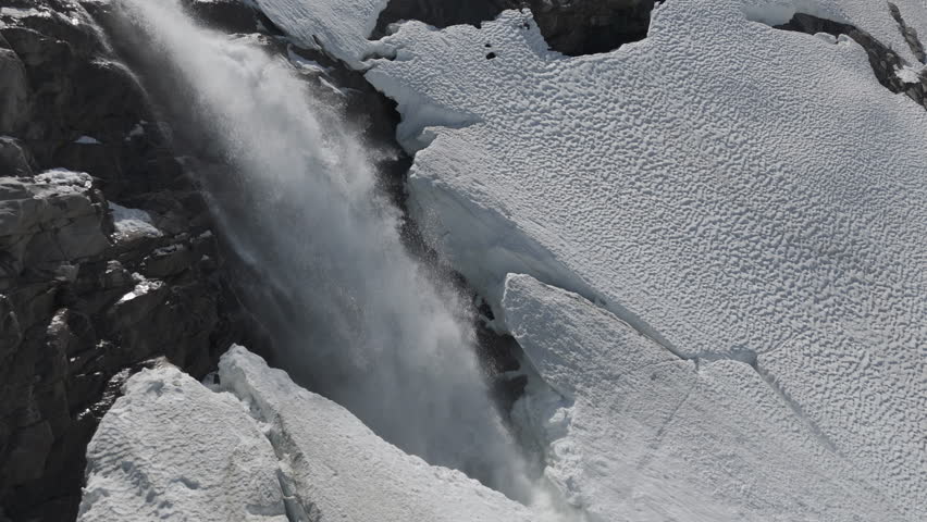Slowmotion drone shot flying around a powerfull waterfall near Langvatnet Lake in Norway close to the Strynefjellsveg breaking through ice and snow on a sunny day surrounded by shiny rocks LOG