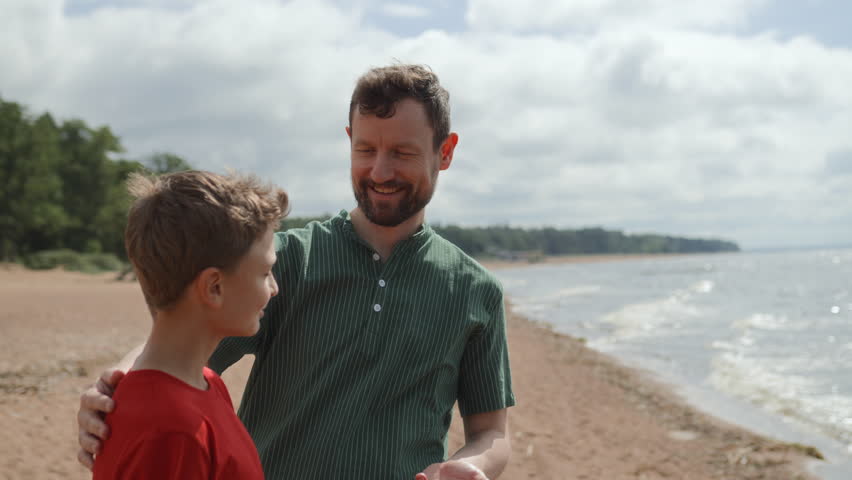 Father and teenage son take leisure walking along coastline of bay. Talking and sharing stories and jokes, smiling and enjoying togetherness. They stop to look at skyline. Family bonding