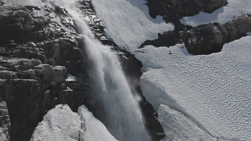 Slowmotion drone shot flying around a powerfull waterfall near Langvatnet Lake in Norway close to the Strynefjellsveg breaking through ice and snow on a sunny day surrounded by shiny rocks LOG