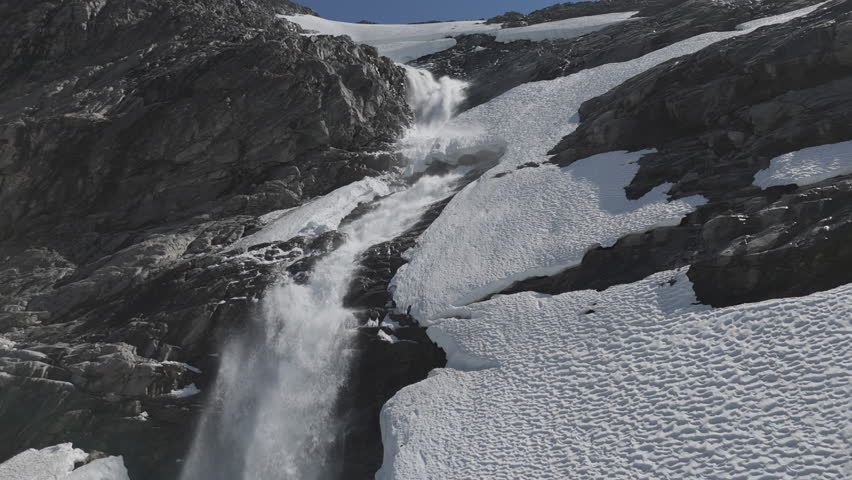 Slowmotion drone shot flying around a powerfull waterfall near Langvatnet Lake in Norway close to the Strynefjellsveg breaking through ice and snow on a sunny day surrounded by shiny rocks LOG