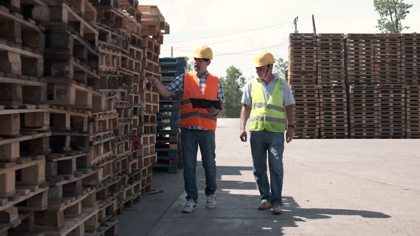 Two workers, walk through the factory yard, checking product numbers and quality. They discuss production details to ensure standards are met.	
