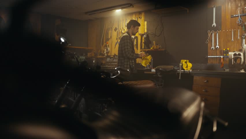 Side view of a guy mechanic in a checkered shirt looking around and cleaning his motorcycle in the workshop