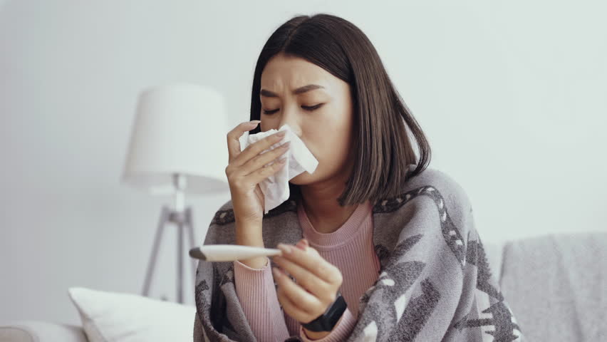 Seasonal flu. Upset asian woman looking at thermometer with fever result, blowing nose and being worried about coronavirus symptoms, slow motion