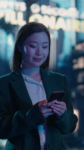 Vertical Screen: Young Asian Woman Using Smartphone while Standing on a Busy City Street at Night, Surrounded by Tall Buildings and Pedestrians. Female Browsing Internet and Checking Social Media