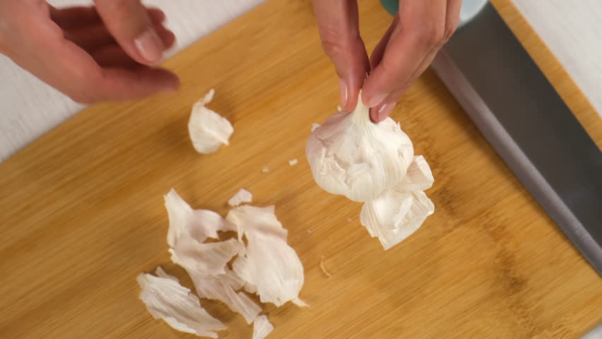 Woman cooking peeling garlic using knife on wooden board, hands close-up. Preparing food, meal on kitchen at home. Cooking dish. Culinary, cuisine, cook with fresh products, ingredients concept.