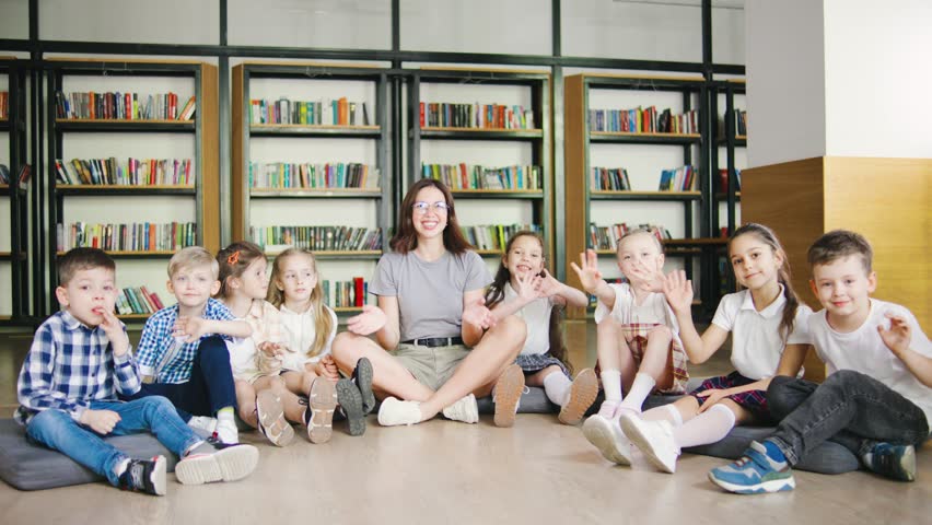 Smiling elementary school kids and their teacher wave at the camera while sitting on cushions in a cozy school library, surrounded by books, sharing a friendly greeting or goodbye