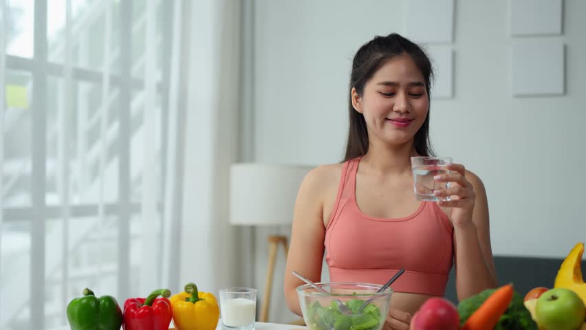 Beautiful Asian woman in sportswear drinking water, vegetable salad, healthy food after exercise on sofa at home. Ideas for taking care of your health in the home kitchen, keeping your body in shape.