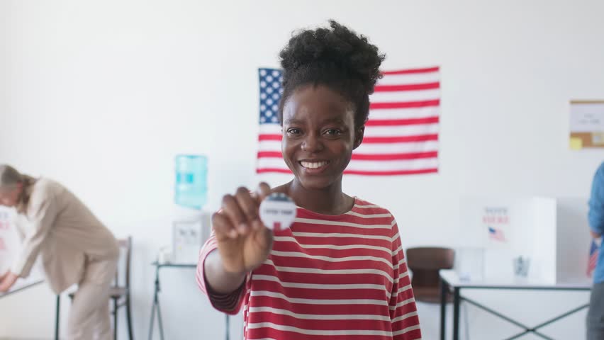 Charming African American Woman with wide smile standing in front of camera. Holding round pin stating girl already voted today. Camera changing focus. In blurred background people filling ballots.