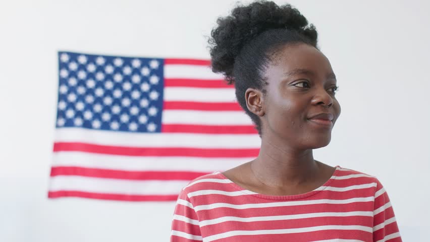 View from below of African American girl standing in front of big American flag hanging on wall. Woman glancing at camera and smiling with joy. Poll station worker proud of work completed.