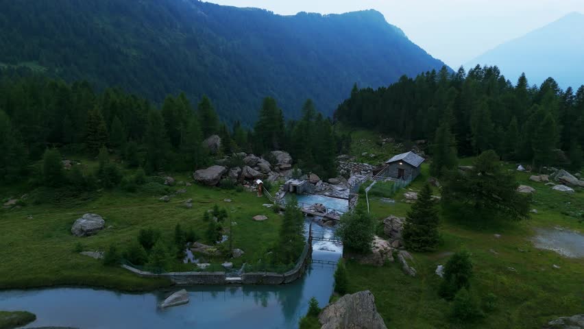 Aerial drone footage over Alps Valmasino, Preda Rossa, Italy, Picturesque mountain cabin nestled in lush green forests with a serene stream running through the alpine landscape. Ascent with tilt down