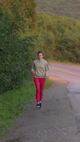 Woman sportsperson in exercise clothes running along country road, taking pleasure in fitness activity. Athletic woman is enjoying outdoor sports training and feeling great on summer. Vertical video