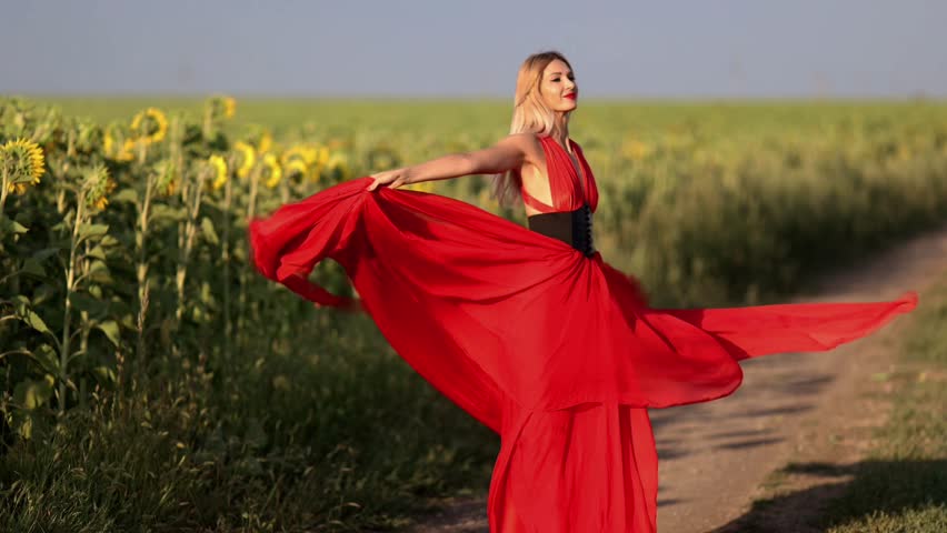 A young woman in a red dress walks through a field of sunflowers.
