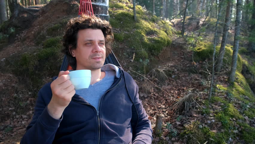 Portrait curly man relaxing lying in hammock drinking tea in forest on nature. Resting relaxed brunet male enjoying time in camping in woodland. Tourist, traveler, camper contemplating outdoors.