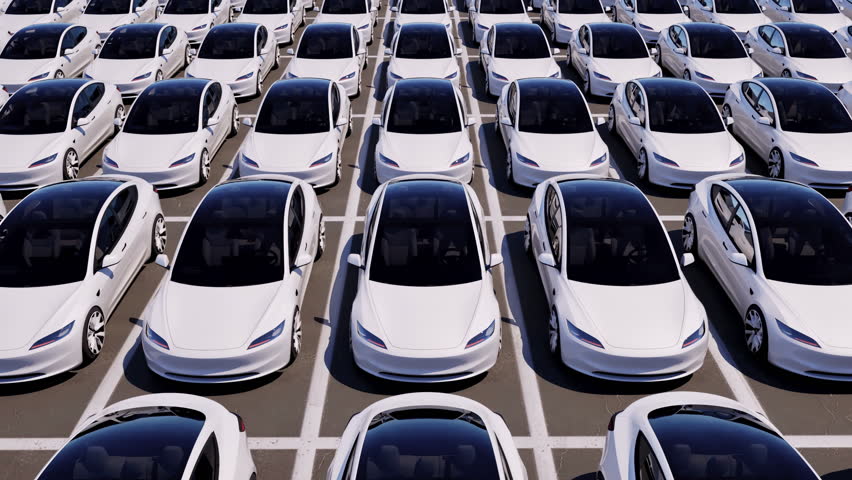 An aerial view over a vast parking lot filled with unsold new white electric vehicles.