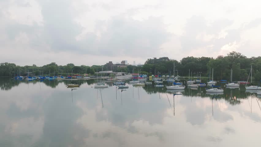 Moving drone view of lake Bde Maka Ska Minneapolis rise to see skyline during daytime in USA. Sailboats in foreground.