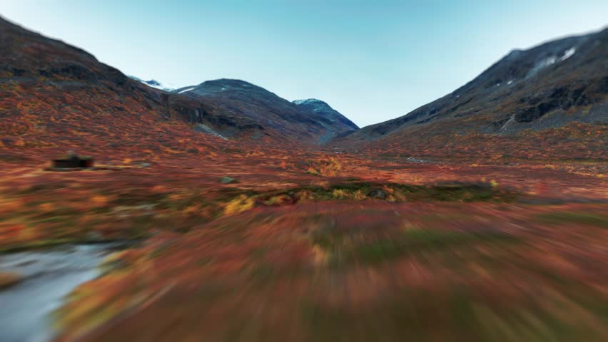 An expansive autumn landscape above the Trollstigen, with vibrant orange and red foliage, contrasted by the distant snowy mountains. Aerial view, fast flight forward.