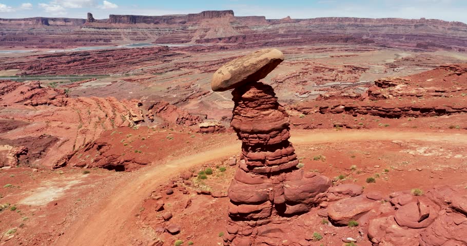 An orbital drone shot of the extremely unique desert landscape of Moab, Utah. The camera rotates around a boulder balancing on top of an isolated rock tower, with intense cliff drops in the background