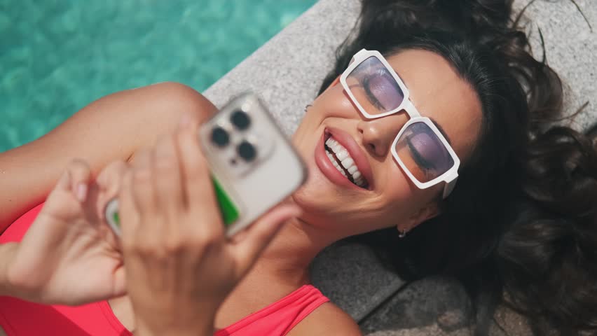 Beautiful girl in pink swimsuit lying on the edge of the pool with smartphone in hand. Tourist using mobile phone in the pool. Relax in luxury pool
