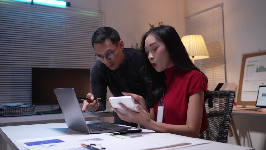 Two asian office workers staying late in the office, sharing a digital tablet and smiling. Happiness radiates as they celebrate their achievements together