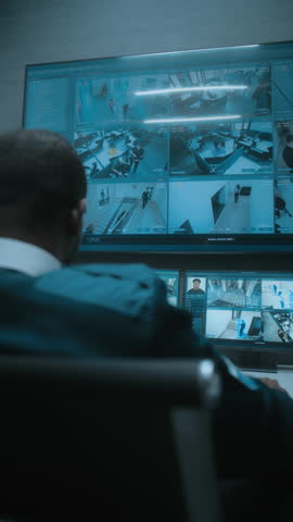 African American male surveillance operator works with security cameras on computer in modern monitoring center. CCTV footage on multiple monitors and big digital screen. Back view. Vertical shot.