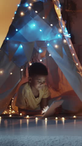 Vertical shot of little boy playing on tablet while lying in teepee decorated with lights in his room