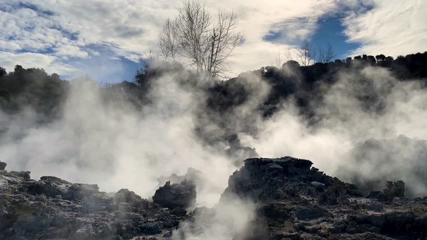 Geothermal steam rising from volcanic landscape on partly cloudy day with blue sky