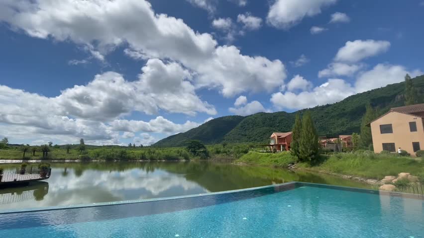 A scenic view of a luxurious resort featuring an infinity pool overlooking a serene lake, with mountains in the background and a bright, cloud-filled sky.