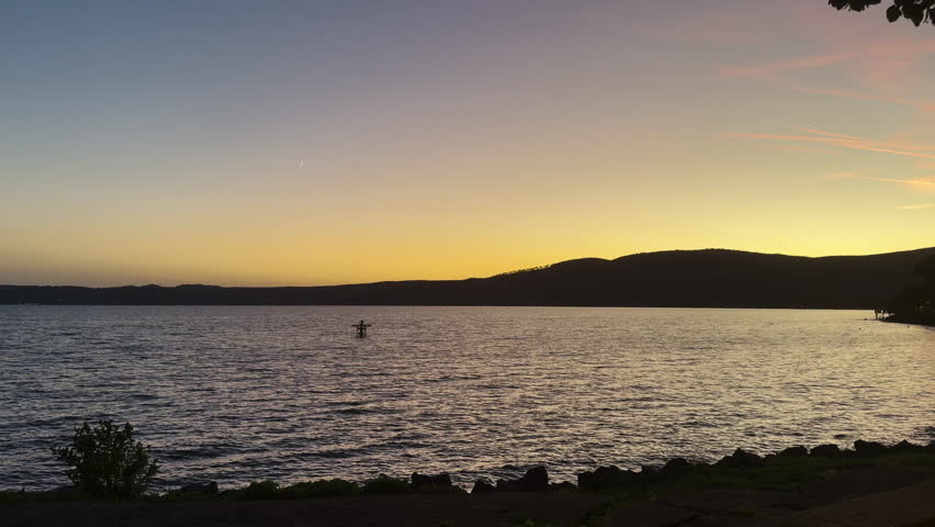 A breathtaking peaceful sunset over Bracciano Lake in Trevignano Romano, with a crescent moon visible above the hills, enhancing the calm and serene atmosphere.