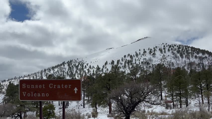 Sunset Crater Volcano National Monument on a partly cloudy day, covered in snow. There is a sign in the foreground. The volcano is a cinder cone located north of Flagstaff in Arizona - USA