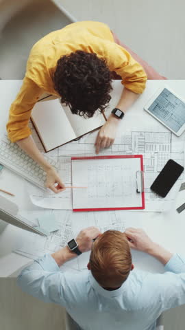 Vertical directly above shot of female architect working on construction plan using tablet, then greeting male colleague with fist bump and discussing drawing together