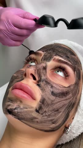 Beautician doing hardware carbon peeling to a young beautiful client in a headdress in goggles lying on a sofa in a beauty salon. Facial cleansing concept. Cosmetology services
