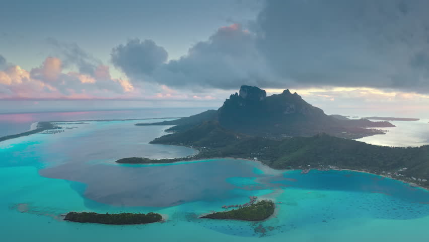 Bora Bora turquoise blue lagoon and green Otemanu mountain in soft sunset light. Coral barrier reef motu under dramatic clouds. Remote wild nature paradise, exotic luxury travel. Aerial drone flight