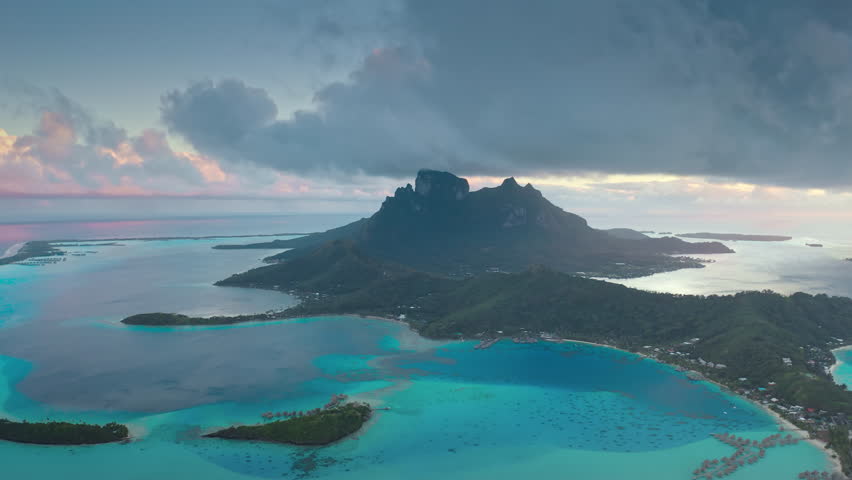 Bora Bora turquoise blue lagoon and green Otemanu mountain in soft sunset light. Coral barrier reef motu under dramatic clouds. Remote wild nature paradise, exotic luxury travel. Aerial drone flight