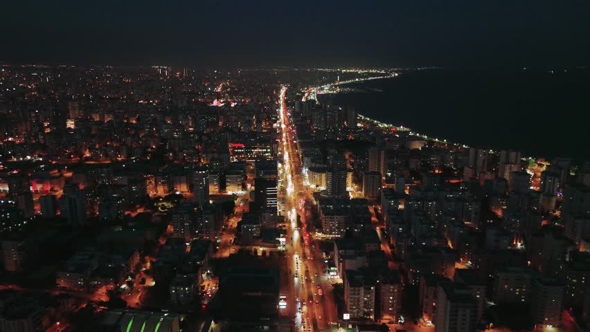 Coastal city with illuminated buildings and streets, bordering the dark mediterranean sea. Cars are driving on the coastal road