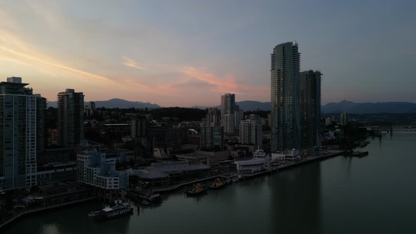 Aerial view of city waterfront and skyscrapers at dusk. Cityscape with backdrop of sunset sky and mountains beyond. Water reflects twilight colors. Serene, urban atmosphere. British Columbia, Canada.