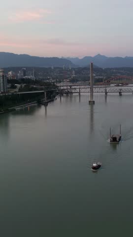 Drone footage of a tugboat pushing a barge down a river at sunset. City skyline and mountains in the background. British Columbia, Canada.