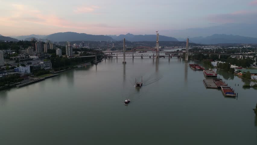 Aerial view of a tugboat pushing a barge along a river at sunset, with a city and mountains in the background. British Columbia, Canada.