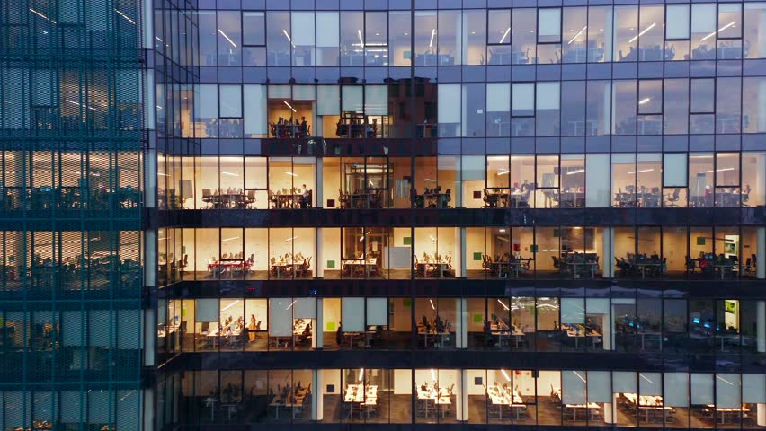This sweeping drone footage captures the illuminated glass skyscraper offices, revealing business professionals hard at work late into the night, highlighting the relentless corporate environment.