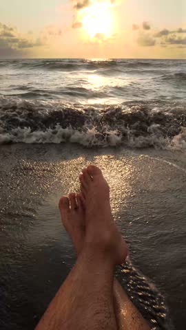 Man relaxing at the sandy beach during sunset, with gentle waves washing over warm feet, creating a calming atmosphere.