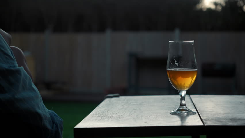 unrecognizable young woman takes a glass of beer from the table while sitting on her terrace overlooking the backyard of her private home in the evening.