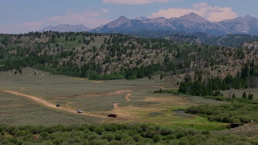 A vast landscape of fields and towering mountains captured from above in Wyoming’s Wind River Range with SUVs driving along dirt road